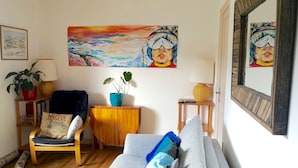 Un salón acogedor con un sofá, una silla, una pequeña mesa de madera, una planta en maceta y un cuadro en la pared.