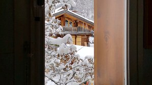 Una ventana con cortina estampada con motivos de corazón, una cabaña de madera con tejado nevado y un paisaje invernal.