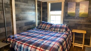 Une chambre avec un mur en bois, un lit recouvert d’une couverture à carreaux, une fenêtre avec des stores et une petite table en bois.