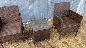 Trois chaises en osier et une table carrée sont disposées sur un sol recouvert de paille, adossées à un mur en métal ondulé.