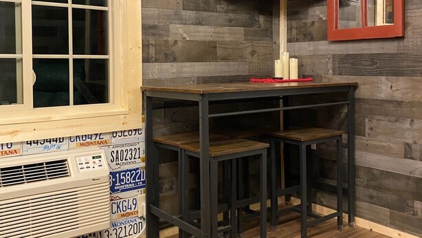 Une pièce avec un plancher en bois, une table et deux chaises, un téléviseur fixé au mur et une fenêtre donnant sur l’extérieur.