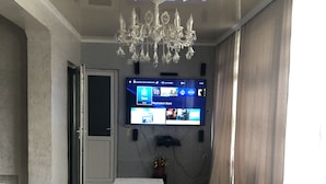 Een kamer met een kroonluchter, een flatscreen televisie aan de muur, een tafel met een witte tafelkleed en een kamerkleed met een patroon.
