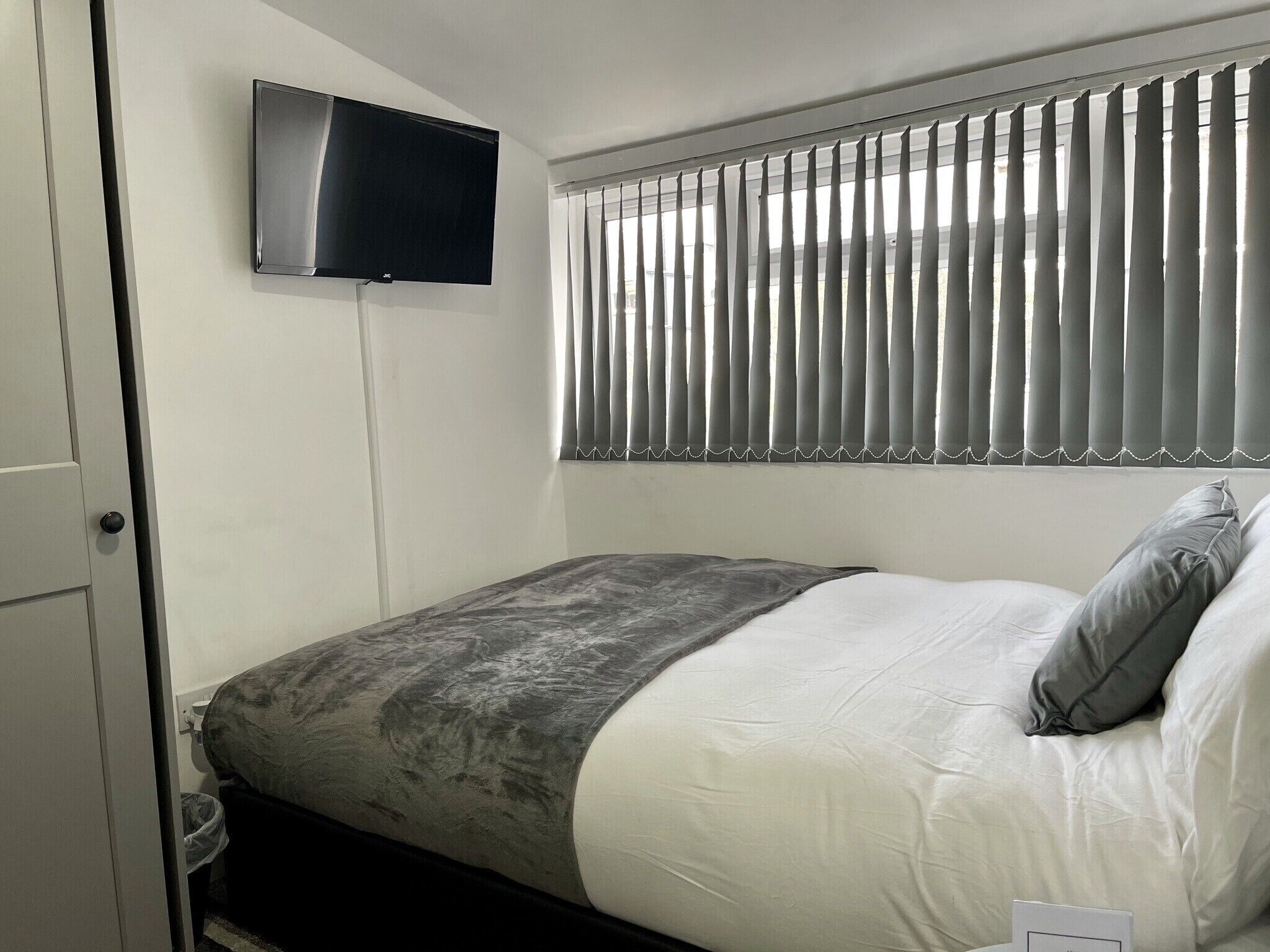 A bedroom with a bed, a television mounted on the wall, and a window with vertical blinds.