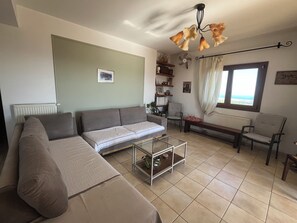 Un salon comprenant un canapé, une table basse en verre, un fauteuil, un banc, un ventilateur de plafond et une fenêtre avec des rideaux.