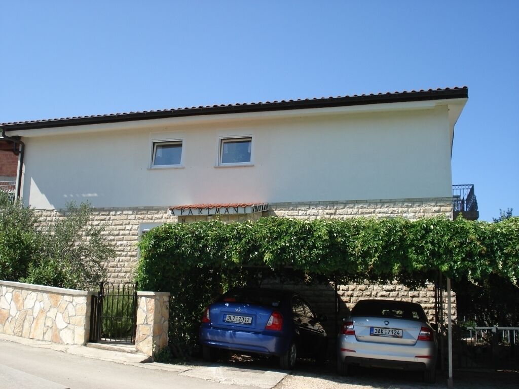 A two-story building with a stone facade and a tiled roof. There are two cars parked in front, and a gate is visible on the left side.