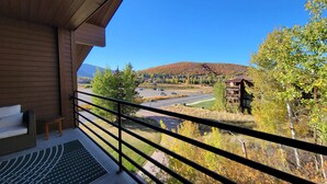 A balcony with a view of a parking lot, a building with a wooden facade, and a hilly landscape in the distance.