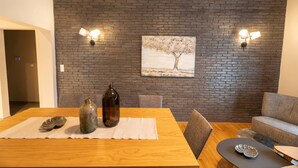 Een eethoek met een bakstenen muur, twee wandlampen, een schilderij van een boom, en een tafel met een placemat, een fles en een kom.