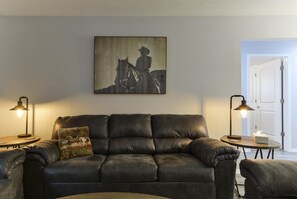 A dark leather sofa with a mounted painting of a horse and rider, two floor lamps, and a small table with a tissue box. - GlamRus Digs (Branson)