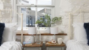 A bedroom with a large window, a bedside table with a lamp, a vase with green plants, and a bottle of water. - Traditional cosy cottage which lies in the heart of St Agnes (St Agnes)