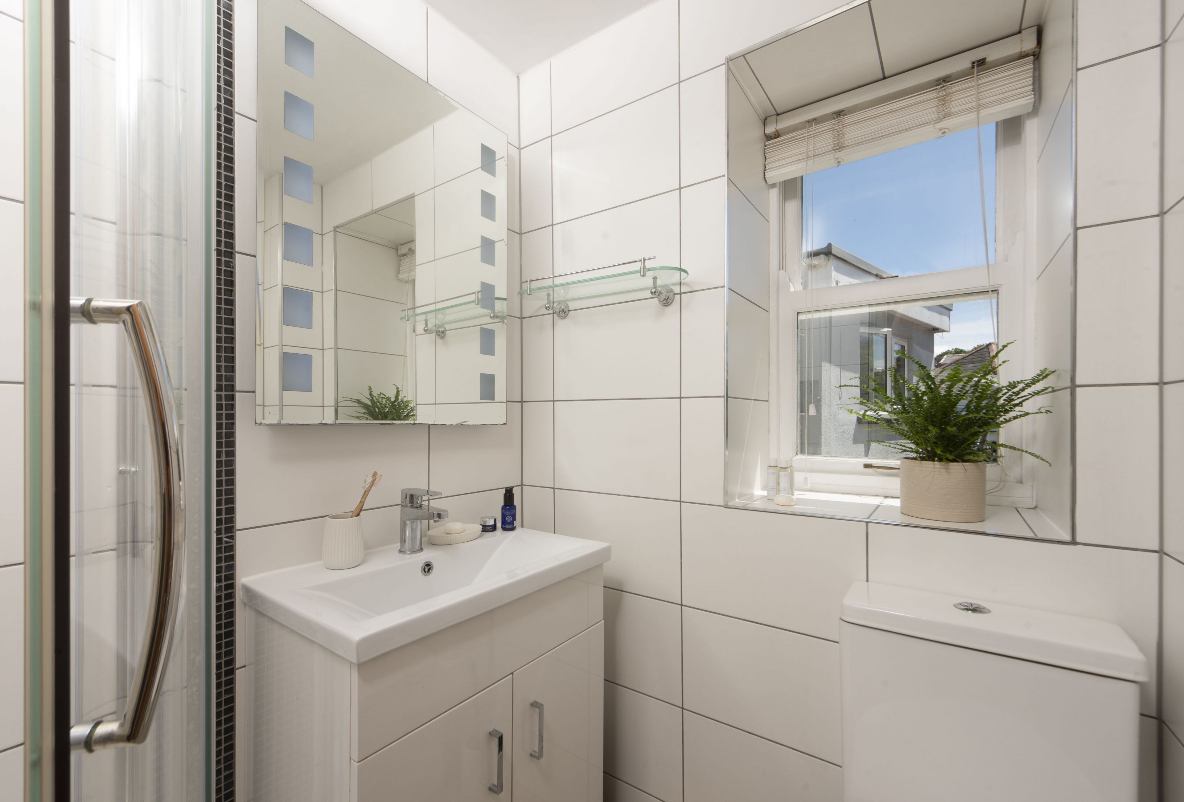 A modern bathroom with a glass shower enclosure, a white sink with a mirror above it, and a window with a potted plant.