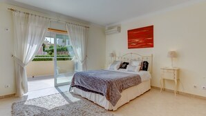 A bedroom with a bed, a nightstand, a lamp, a red abstract painting, and a view of greenery outside.