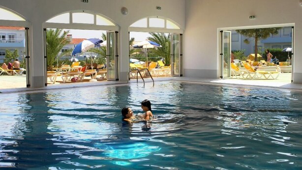 An indoor swimming pool with clear glass walls, a wooden ceiling, and sun loungers outside.