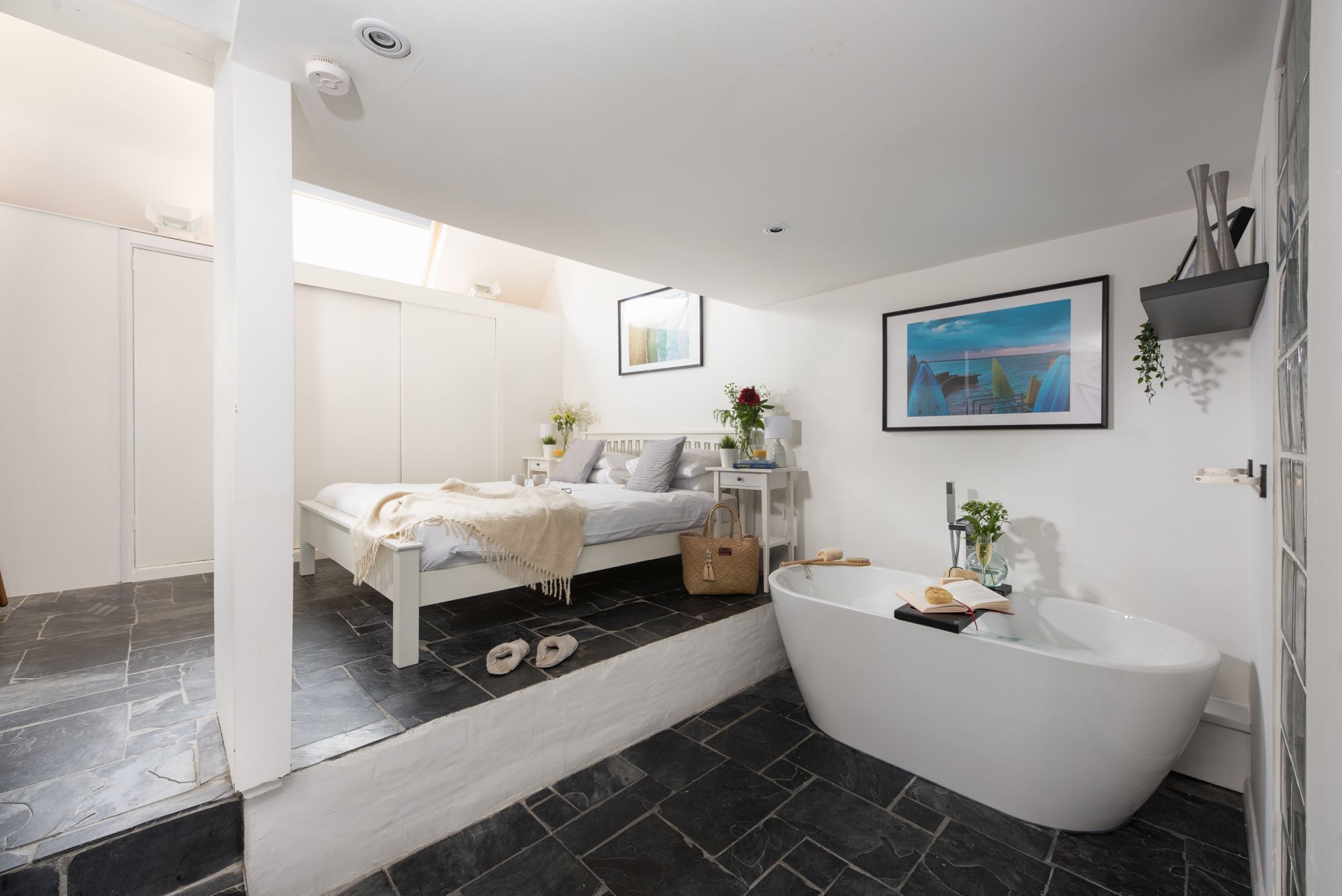 A modern bathroom with a bathtub, a shower, and a small bedside table with a lamp and a plant.