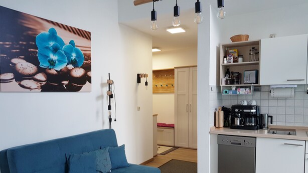 Ett modernt vardagsrum med en blå soffa, ett soffbord och en köksdel med vita skåp och vitvaror.