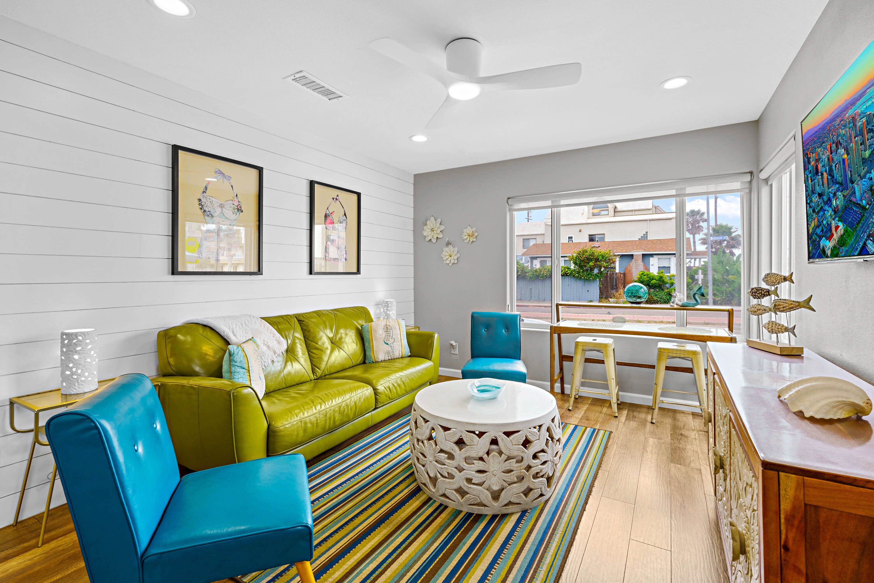 A modern living room with a green sofa, blue chairs, a round coffee table, and a large window with a view of buildings.