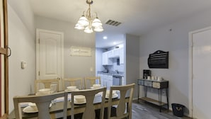 A dining area with a wooden table and chairs, a chandelier, and a kitchen in the background.