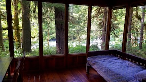 A cabin with a wooden floor, a bed with a blue quilt, and large windows offering a view of a forest.