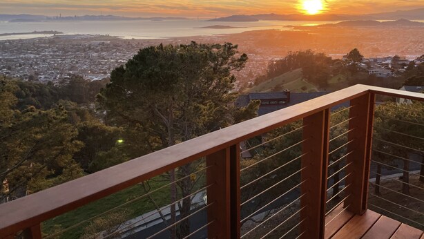 Un balcon offrant une vue sur un coucher de soleil au-dessus d’une ville, avec un pont et une étendue d’eau visibles.