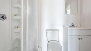 A white bathroom with a toilet, a sink, and a mirror. There are shelves with toiletries.