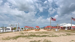 Zona de playa con mesas de picnic y sombrillas rojas, un puerto deportivo con barcos y banderas estadounidenses.