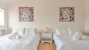 A hotel room with two beds, white bedding, a nightstand with a lamp, and two posters on the wall. - Bungalows at Common Ground - Unit 15 Dbl Queen (Montauk)