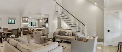 A spacious living area with a high ceiling, a staircase, and a kitchen in the background.