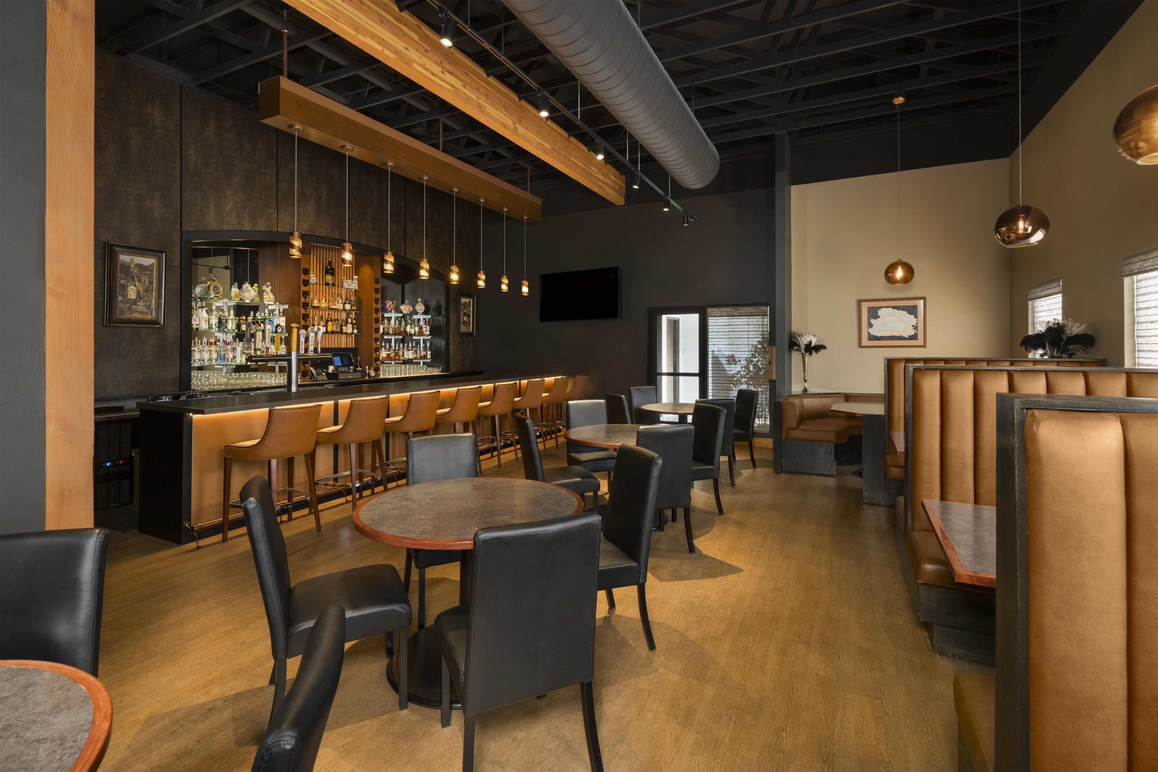Een bar met een toog, krukken en diverse flessen. Er zijn tafels en stoelen klaar voor het diner.