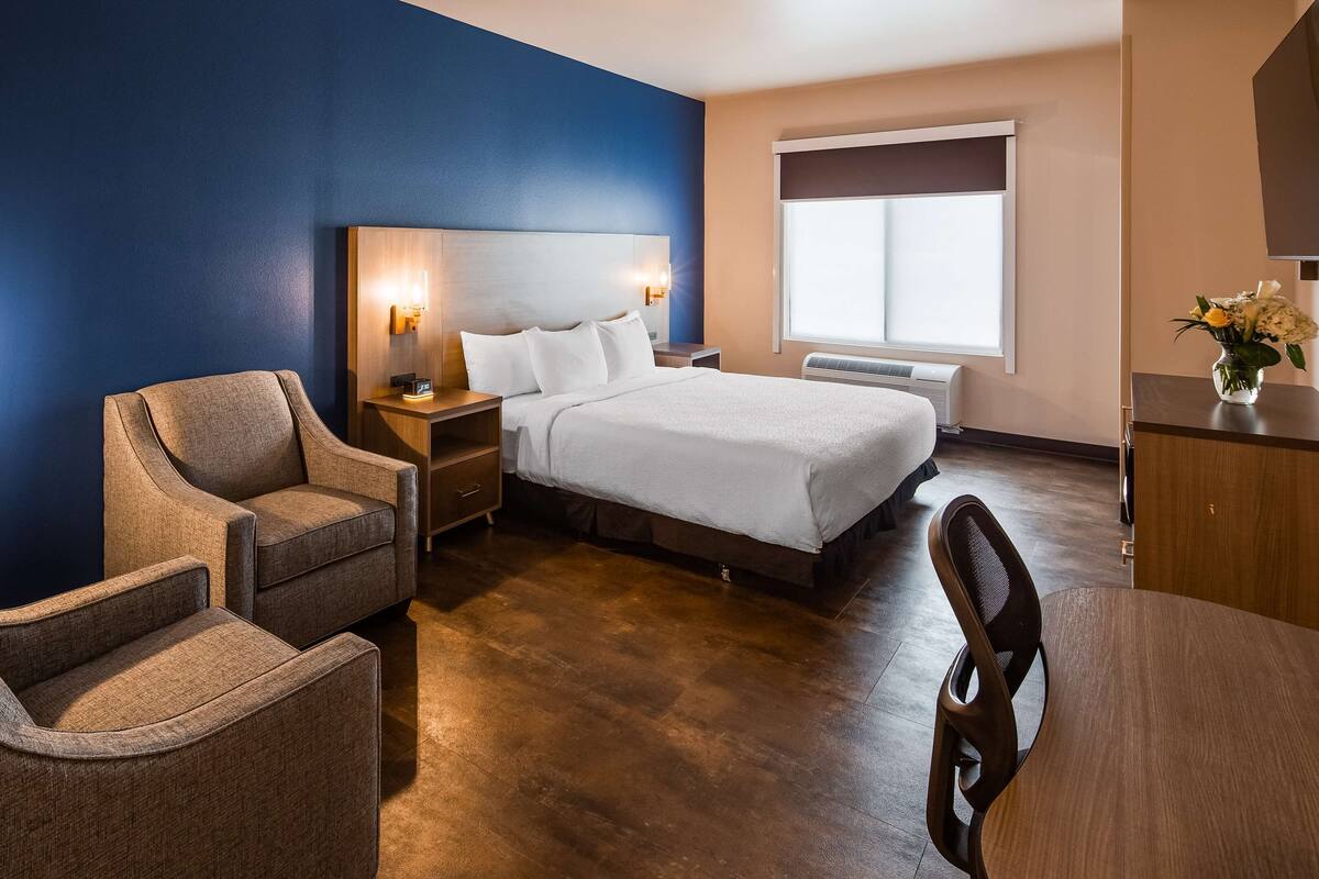 A hotel room with a bed, two armchairs, a nightstand with a lamp, a dresser, and a window with blinds.