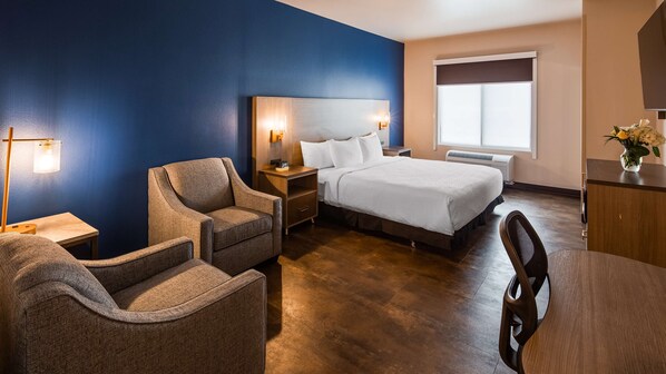 A hotel room with a bed, two armchairs, a nightstand with a lamp, a dresser, and a window with blinds.