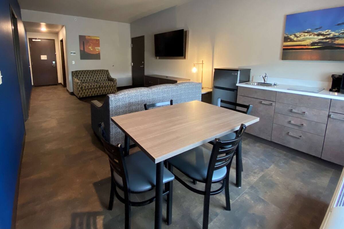 Habitación de hotel compacta con kitchenette, zona de comedor con mesa y sillas, sofá y televisor montado en la pared.