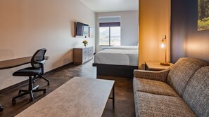 A hotel room with a bed, a desk with a chair, a sofa, a TV mounted on the wall, and a window with a view of the outdoors.