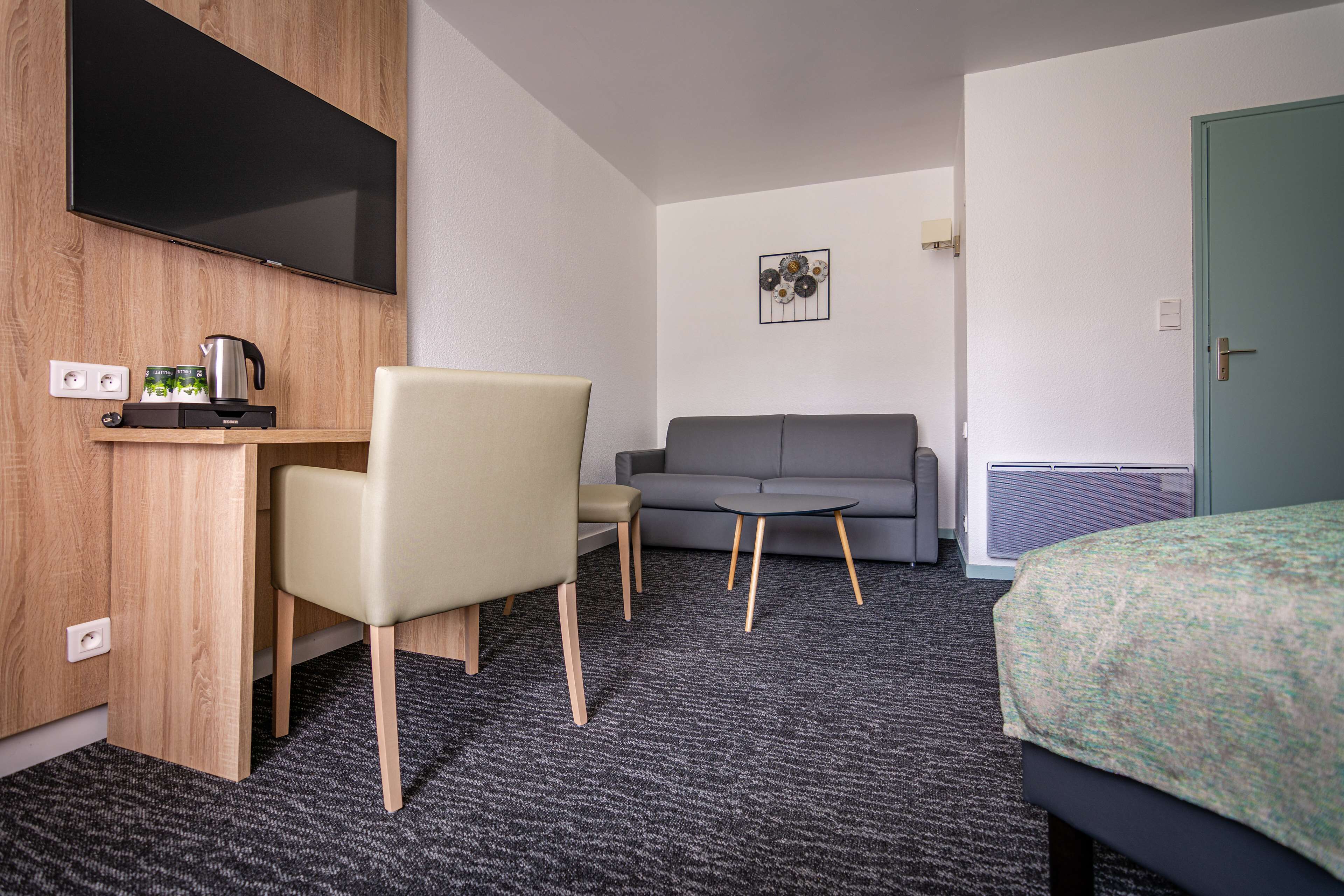 A hotel room with a bed, a desk with a chair, a sofa, a TV mounted on the wall, and a small table.