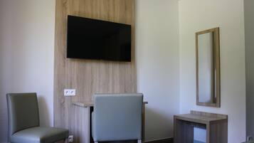 A hotel room with a flat-screen TV mounted on a wooden panel, a small desk with a chair, a bedside table, and a mirror on the wall.