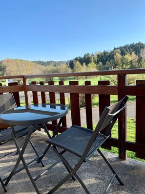 Een balkon met een glazen tafel en twee stoelen, met uitzicht op een groen landschap met bomen en een heuvel.