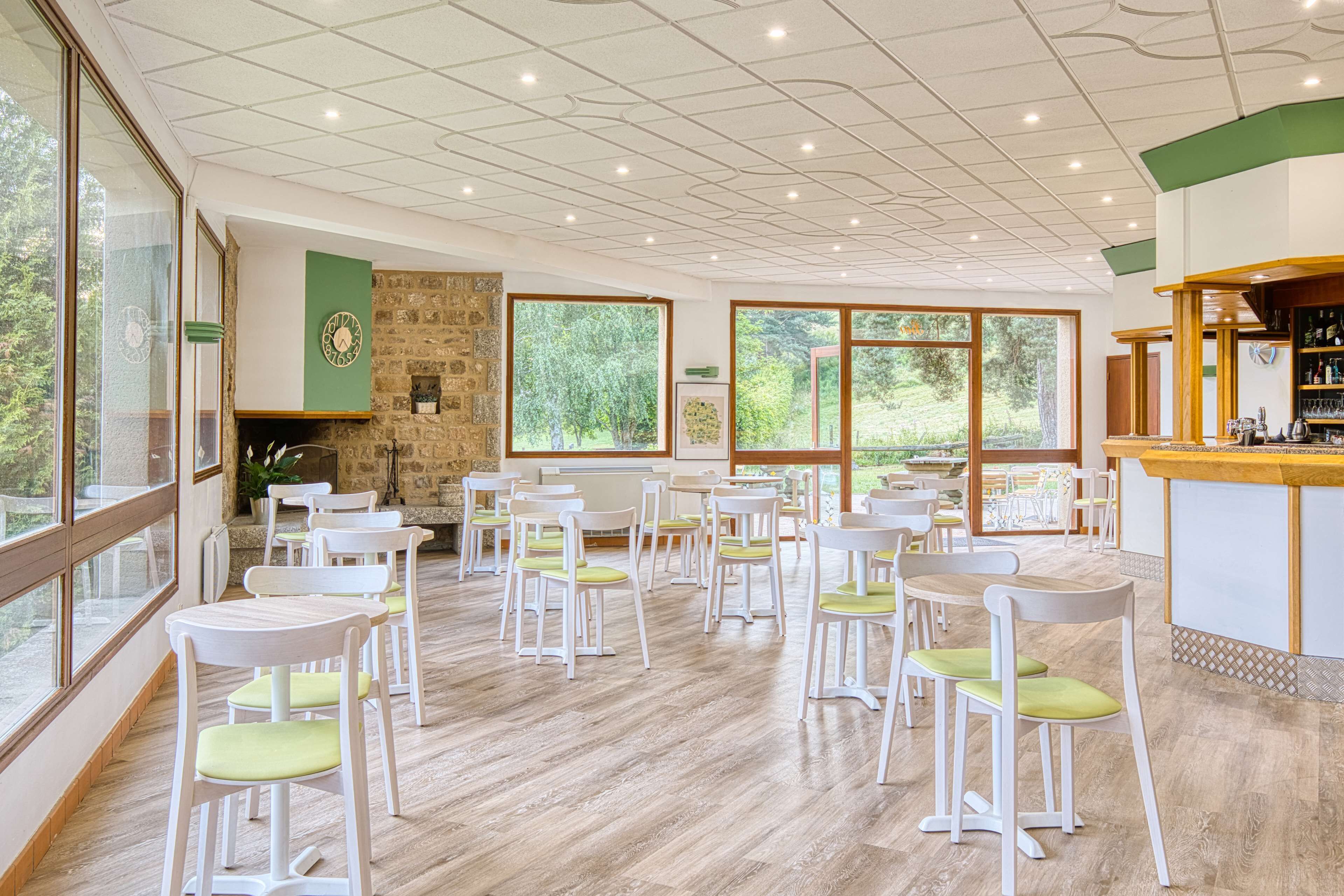 A spacious indoor dining area with white chairs and tables, large windows offering views of greenery, and a stone wall with a clock.