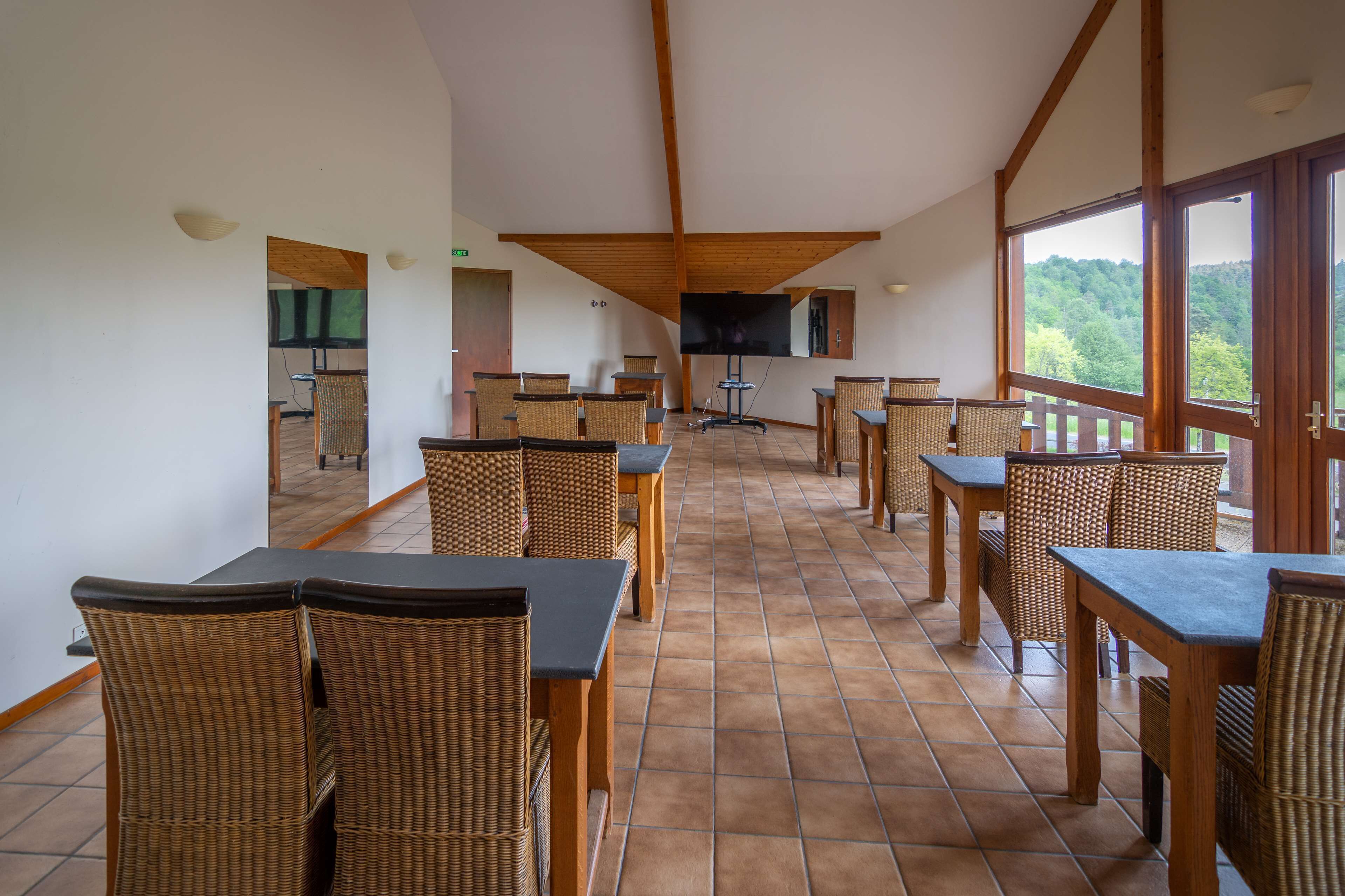 A spacious dining area with high ceilings and large windows, featuring wicker furniture and a flat-screen TV mounted on the wall.