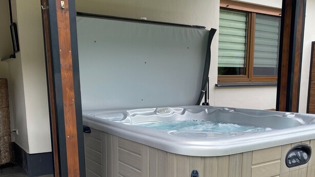 A hot tub with a wooden frame and a control panel, located in an outdoor area with tiled flooring and a window with blinds.