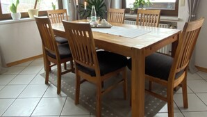 A dining area with a wooden table and chairs, large windows with curtains, and potted plants.