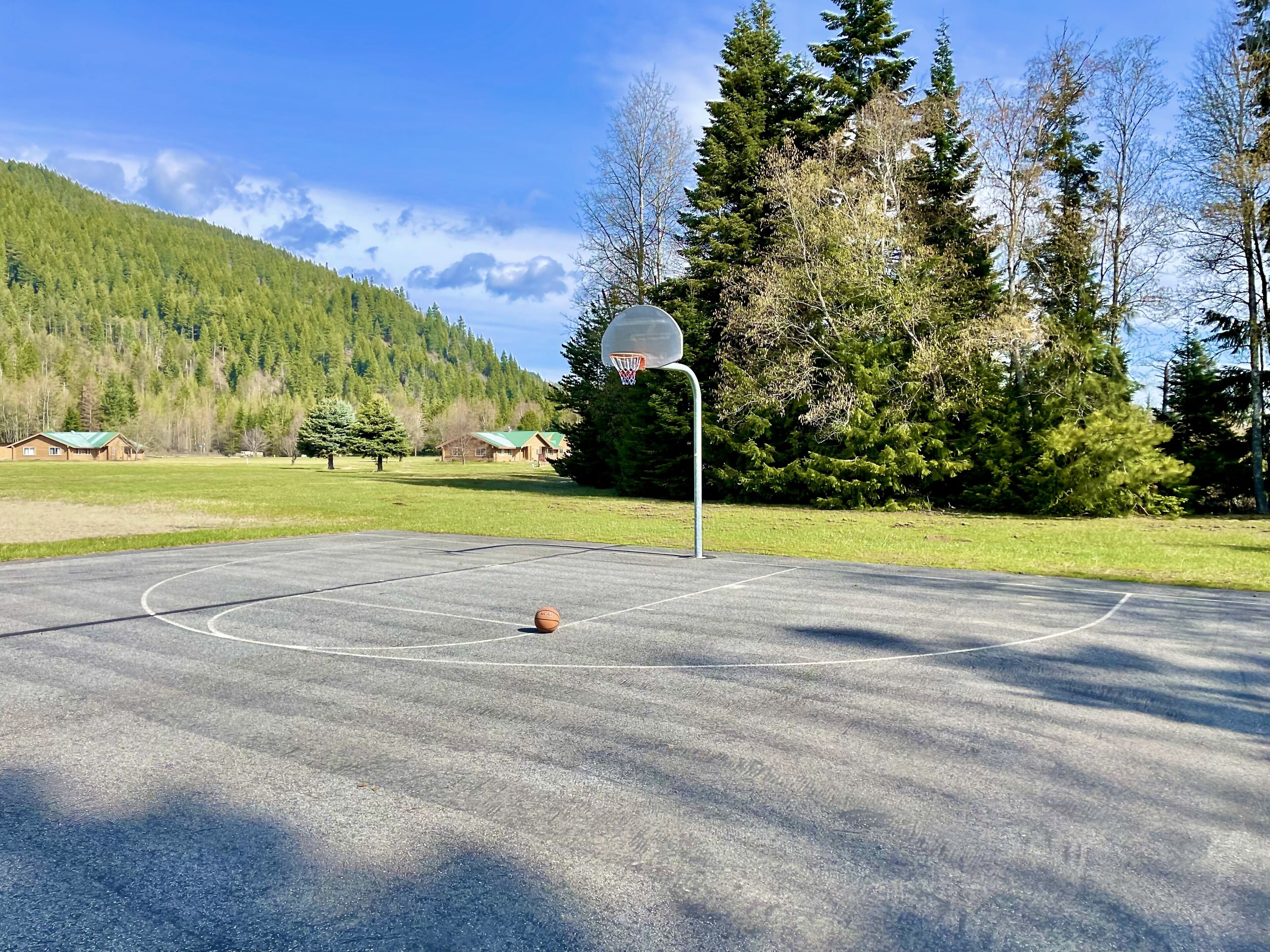 An outdoor basketball court with a hoop, surrounded by trees and a mountain in the background.