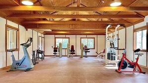 A spacious gym with various exercise equipment, large windows, and wooden beams on the ceiling.