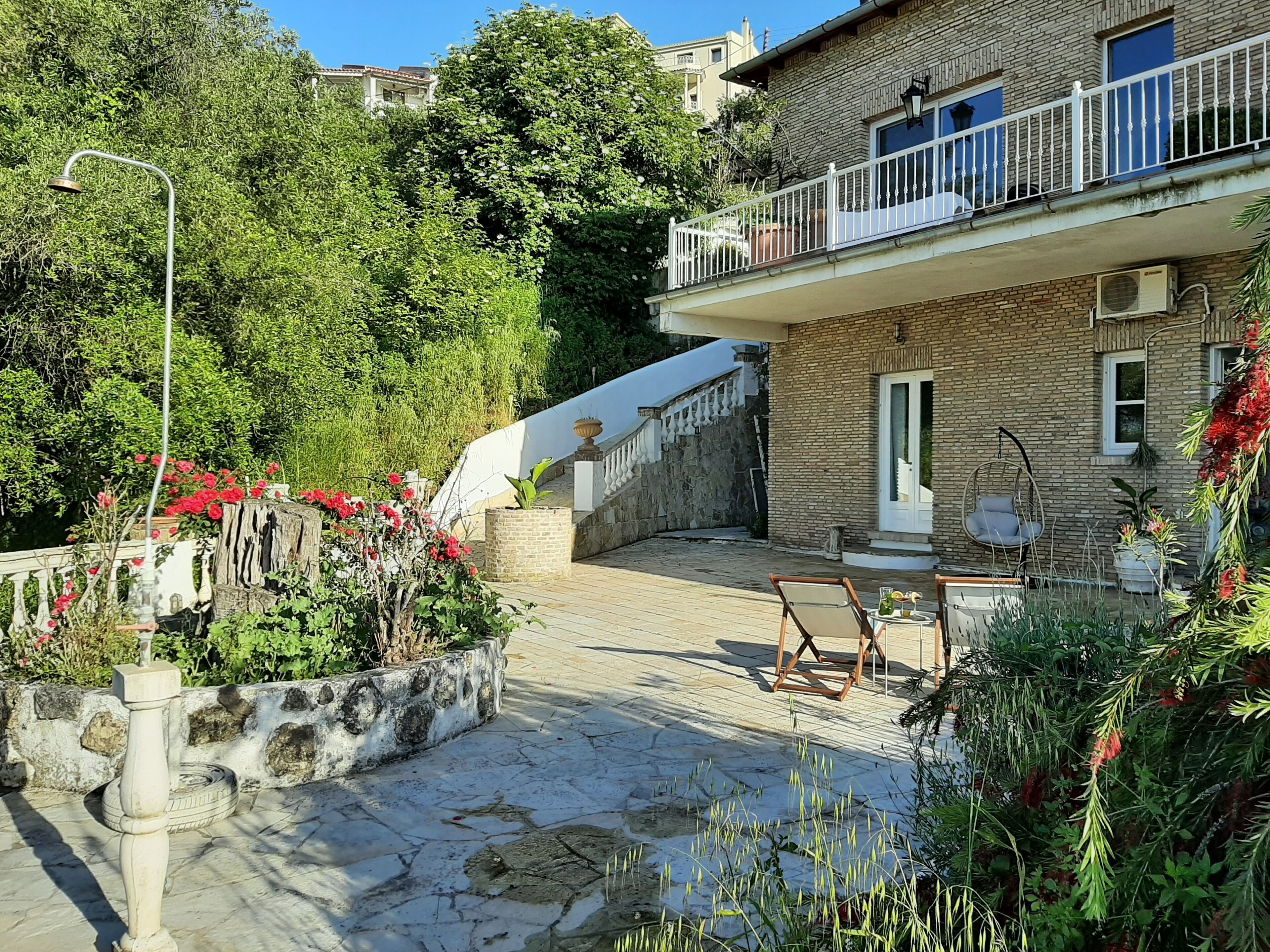 Ein Innenhof mit Steinboden, einer Holzbank und einem Hängesessel. Eine Treppe führt zu einem Balkon mit weißer Reling.