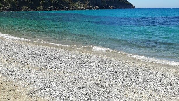 A pebble beach with clear turquoise water and a green hill in the background.