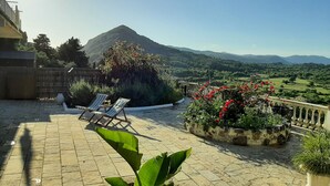 A rooftop terrace with two lounge chairs, a potted plant, and a view of a mountain and green fields.