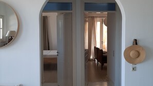 A hallway with an arched doorway leading to a bedroom with a bed and curtains. A mirror and a hat are visible on the walls.