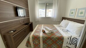 Ein Schlafzimmer mit einem Bett, einem an einem Holz-Kopfteil montierten Fernseher, einem Fenster mit Jalousien und gerahmten Bildern an der Wand.