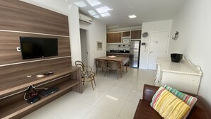 Ein modernes Wohnzimmer mit Flachbildfernseher, einer Holzunterhaltungsecke, einem Essbereich mit Tisch und Stühlen sowie einer Küche im Hintergrund.