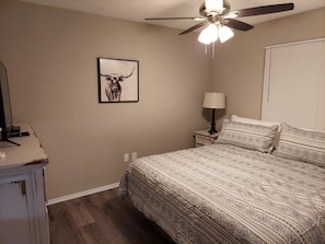 Une chambre à coucher comprenant un lit, une table de chevet, une lampe, un ventilateur de plafond, un tableau accroché au mur et une fenêtre avec des stores.