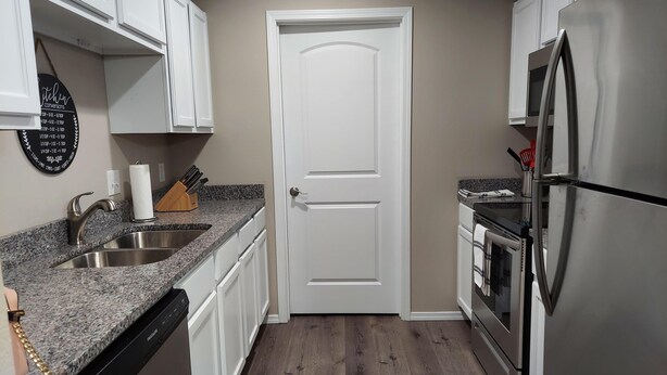 A compact kitchen with white cabinets, a stainless steel refrigerator, a dishwasher, and a granite countertop. - Mystic Ranch Condo (Branson)