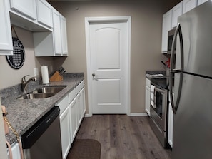 A compact kitchen with white cabinets, a stainless steel refrigerator, a dishwasher, and a granite countertop.