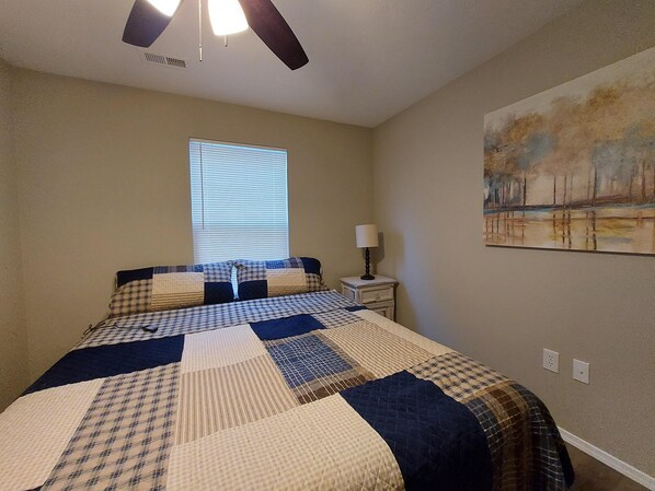 A bedroom with a large bed, a ceiling fan, a window with blinds, a bedside table with a lamp, and a painting on the wall. - Mystic Mountain Condo (Branson)